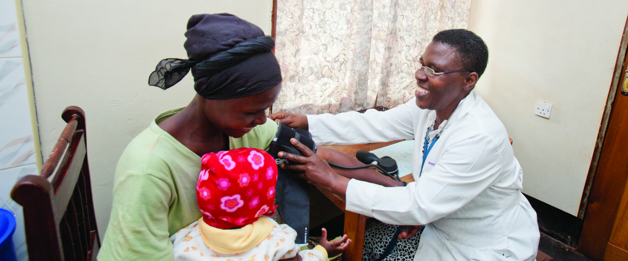 A healthcare worker in a white coat smiles while measuring a woman’s blood pressure; the woman holds a baby dressed in a red and white hat on her lap in a small clinic room.