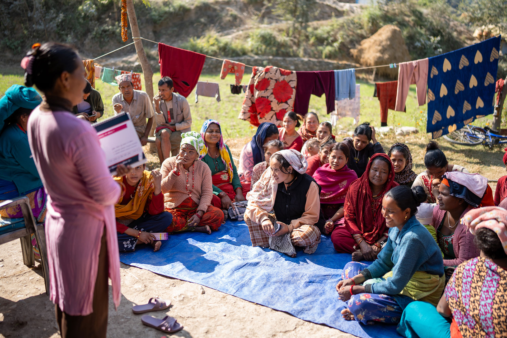 A group of women sit outdoors on a blue mat, attentively listening to a woman standing and holding a booklet. Laundry hangs on clotheslines in the background, and the setting is sunny and rural.