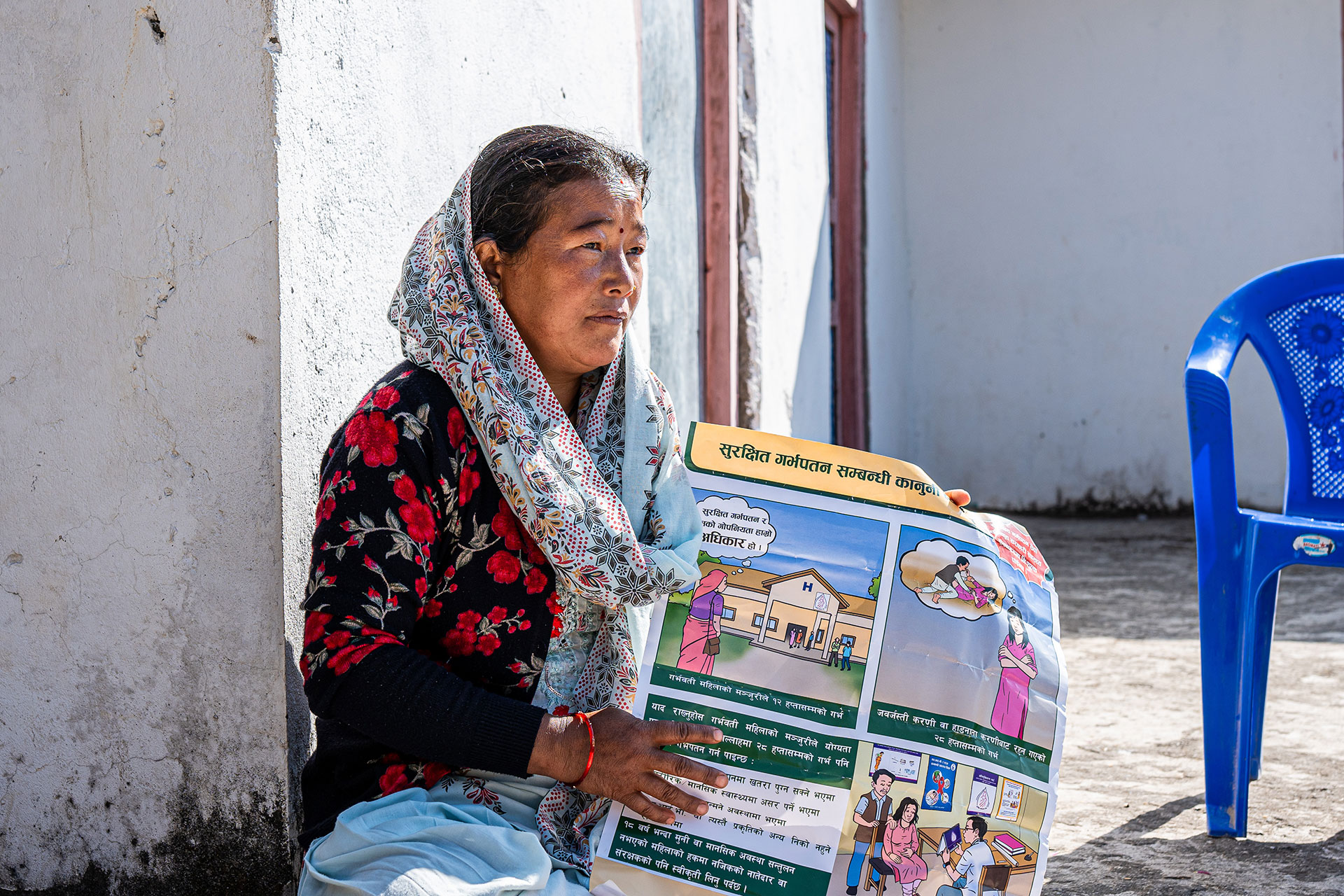 A woman sits on the ground outdoors, holding a colorful illustrated poster with Nepali text. She has a shawl draped over her head and wears a black and red sweater. A blue plastic chair is nearby.