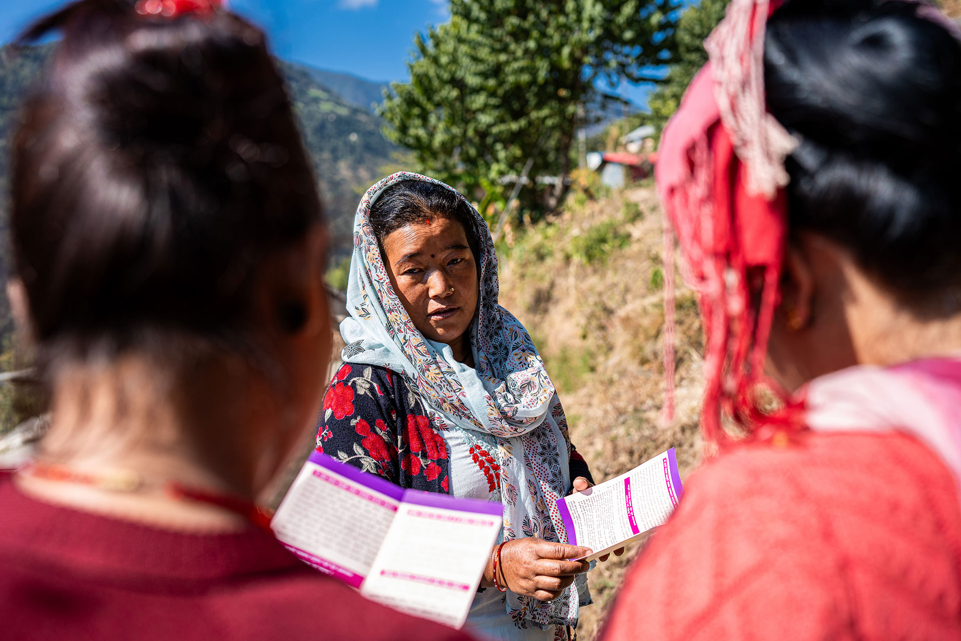 A woman in a floral scarf stands outdoors, holding a pamphlet and talking to two other women with braided hair, who are partially visible from behind. Trees and mountains are in the background under a clear sky.