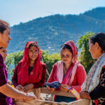 Five women stand together outdoors in a rural, mountainous area, some smiling and wearing colorful traditional clothing. One woman holds an open booklet as they appear to engage in a group discussion. Lush hills are visible in the background.
