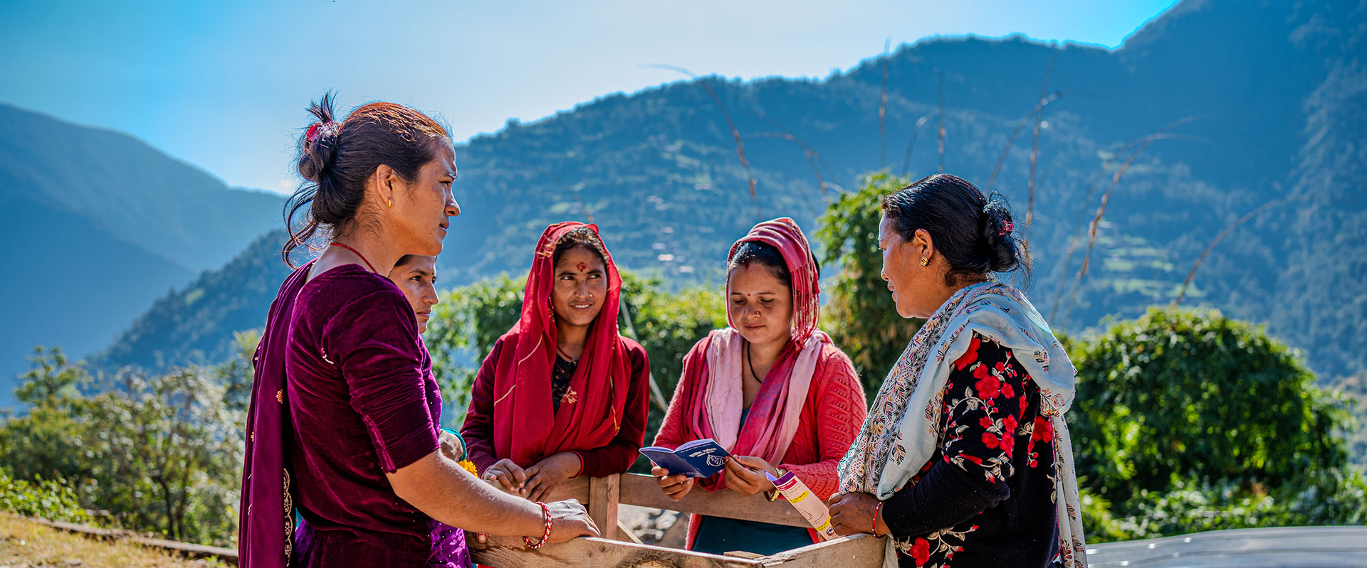 Five women stand together outdoors in a rural, mountainous area, some smiling and wearing colorful traditional clothing. One woman holds an open booklet as they appear to engage in a group discussion. Lush hills are visible in the background.