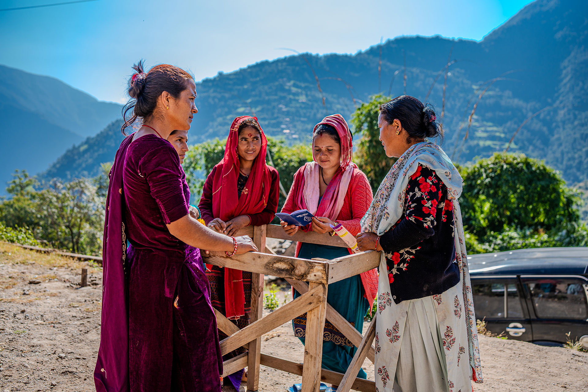 Five women in colorful traditional clothing stand outdoors near a wooden structure, with green mountains and blue sky in the background. One woman holds an open notebook as they engage in conversation.
