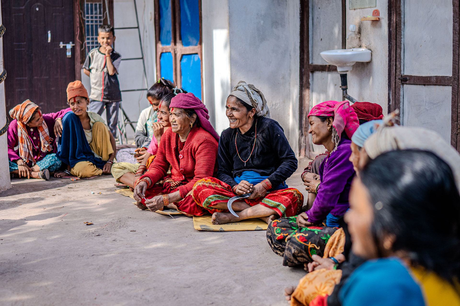 A group of women in colorful traditional clothing sit in a circle outdoors, smiling and talking, while a few children watch nearby. They appear relaxed and engaged in conversation beside a building with blue doors.