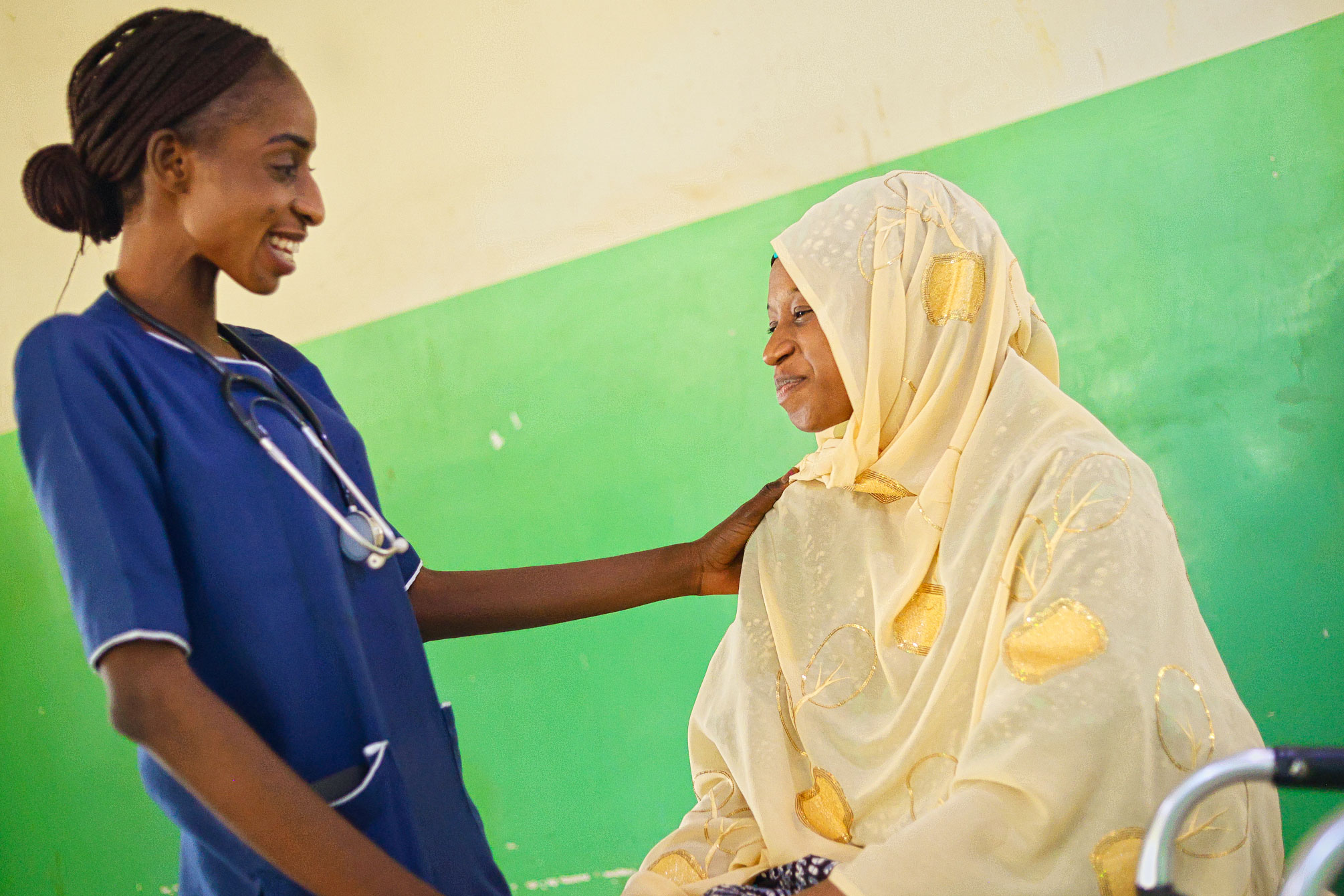 A smiling healthcare worker in blue scrubs gently touches the shoulder of a woman in a cream and gold hijab, both sharing a warm, reassuring moment against a green and beige wall.