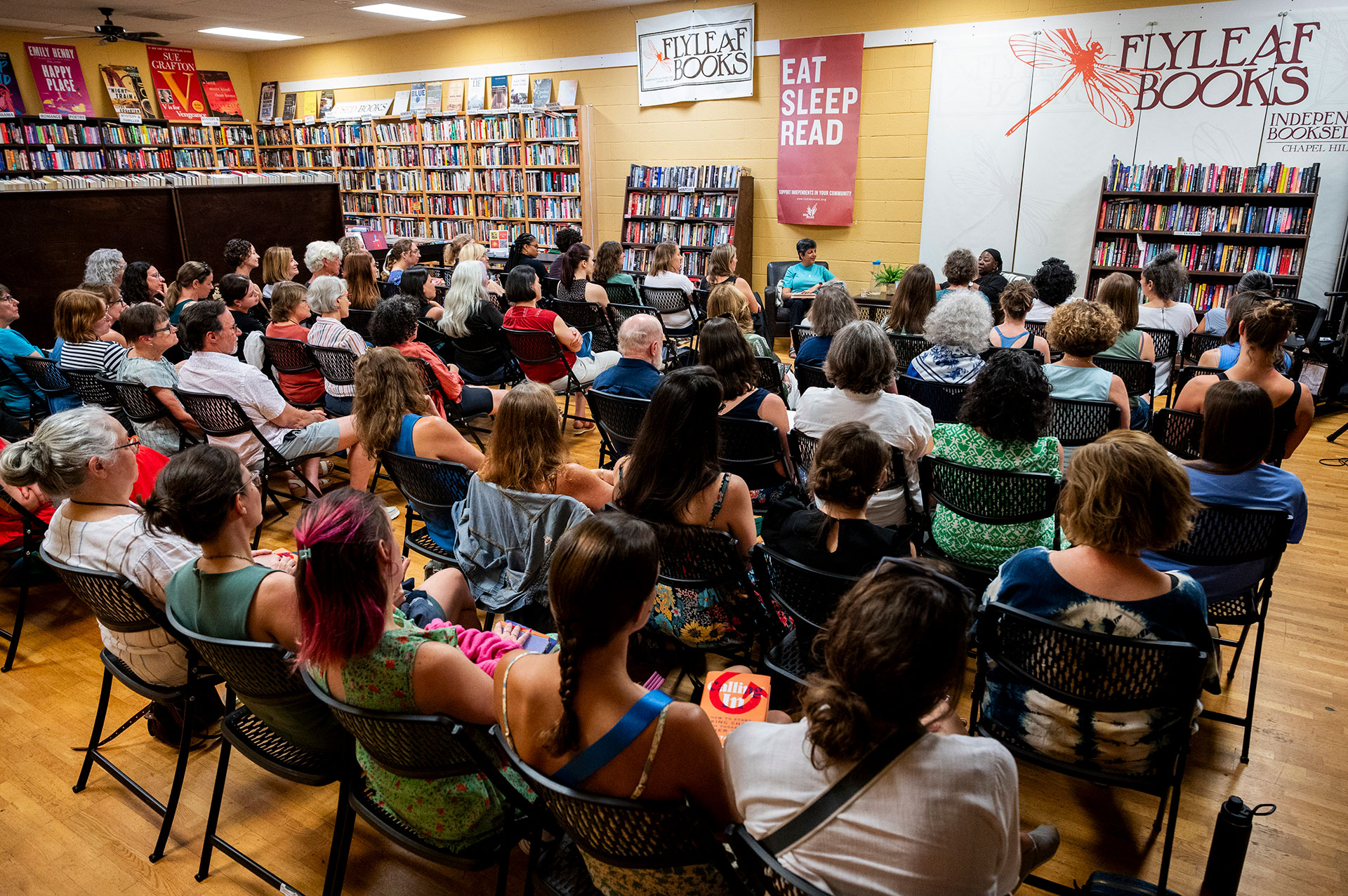 Ipas Read to Resist event with Dr. Loretta Ross A diverse audience sits in rows of chairs at Flyleaf Books, attentively listening to a panel discussion. Bookshelves line the walls, and banners reading "EAT SLEEP READ" and "FLYLEAF BOOKS" are visible at the front.