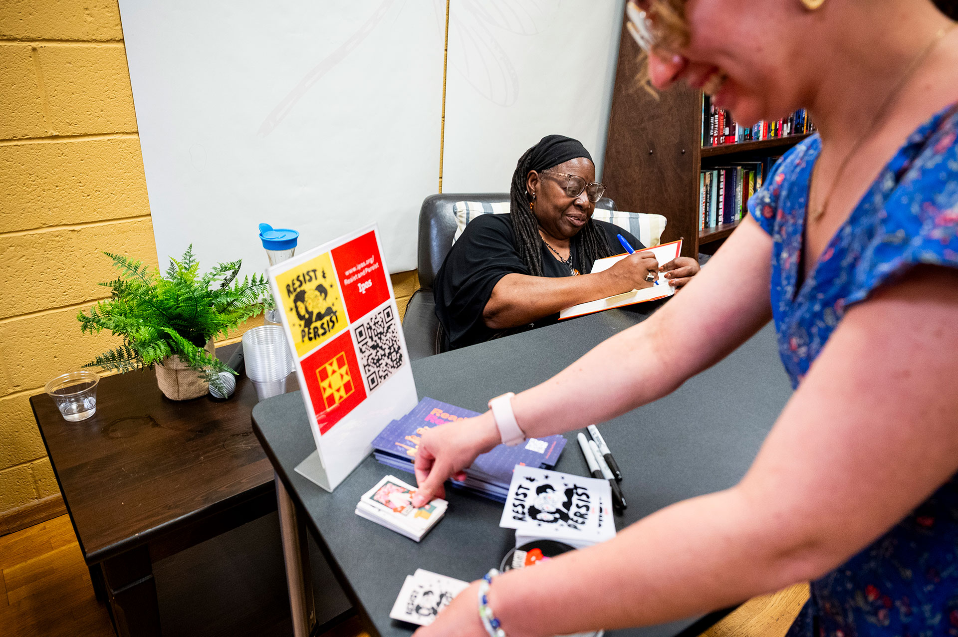 Dr. Loretta Ross sits at a table signing a book, smiling, while another person in the foreground arranges stickers and books on the table. A small plant and water cup sit nearby.