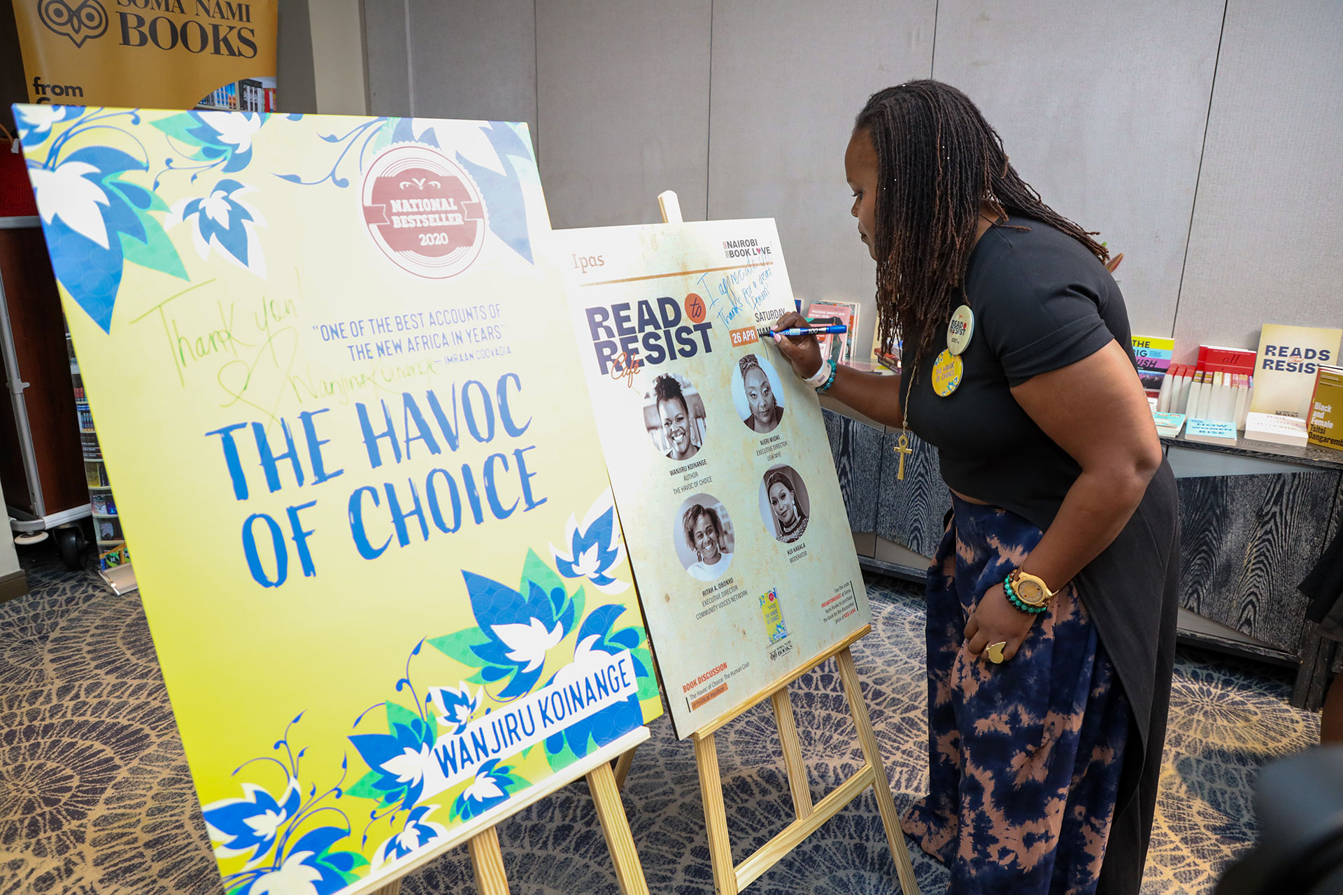 Author, Wanjiru Koinange at the Ipas and Nairobi Book Love Read to Resist Event. A woman signs a poster at a book event. Next to her is a display featuring the cover of "The Havoc of Choice" by Wanjiru Koinange, labeled as an award-winning book. Books and more posters are visible in the background.