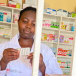 A pharmacist in a white coat examines a pack of pills, including options for postabortion contraception, behind metal bars in front of shelves filled with various medicines and pharmaceutical products.