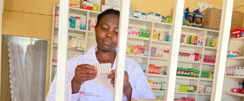 A pharmacist in a white coat examines a pack of pills, including options for postabortion contraception, behind metal bars in front of shelves filled with various medicines and pharmaceutical products.