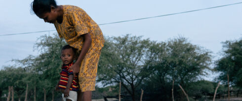 A woman in a yellow patterned dress stands outdoors, bending over a young child in a striped shirt who stands close to her leg. Trees and a fence are visible in the background under a clear sky.