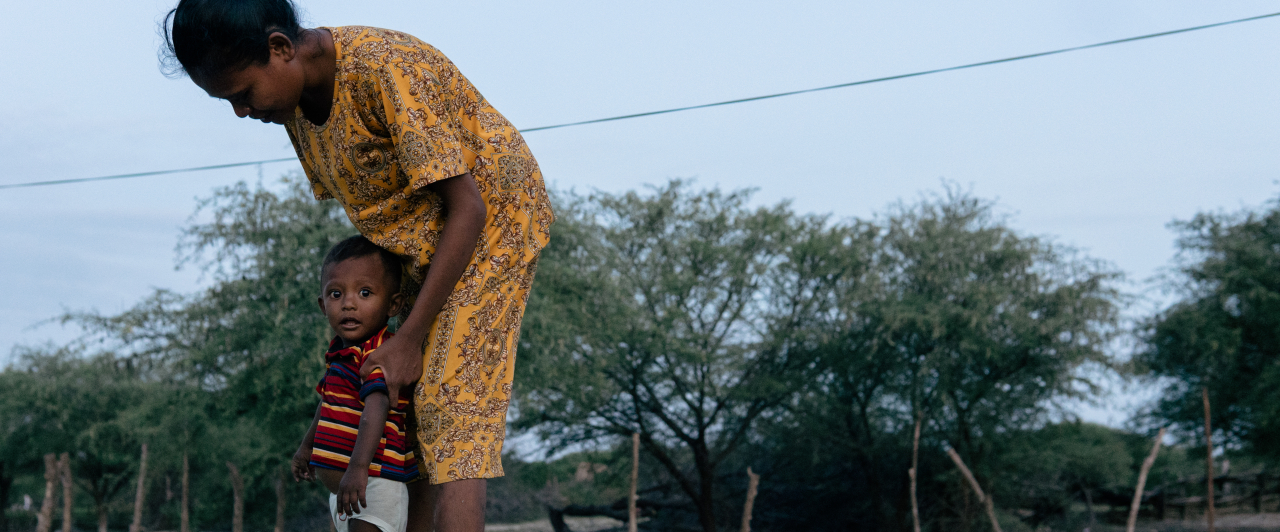 A woman in a yellow patterned dress stands outdoors, bending over a young child in a striped shirt who stands close to her leg. Trees and a fence are visible in the background under a clear sky.