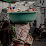 A person with a patterned scarf covering their head carries a green basin filled with plastic bottles on their head, walking through an outdoor area with tents and people in the background—an everyday scene in DRC conflict zones facing reproductive health challenges.