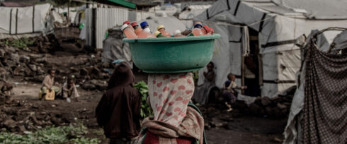 A person with a patterned scarf covering their head carries a green basin filled with plastic bottles on their head, walking through an outdoor area with tents and people in the background—an everyday scene in DRC conflict zones facing reproductive health challenges.