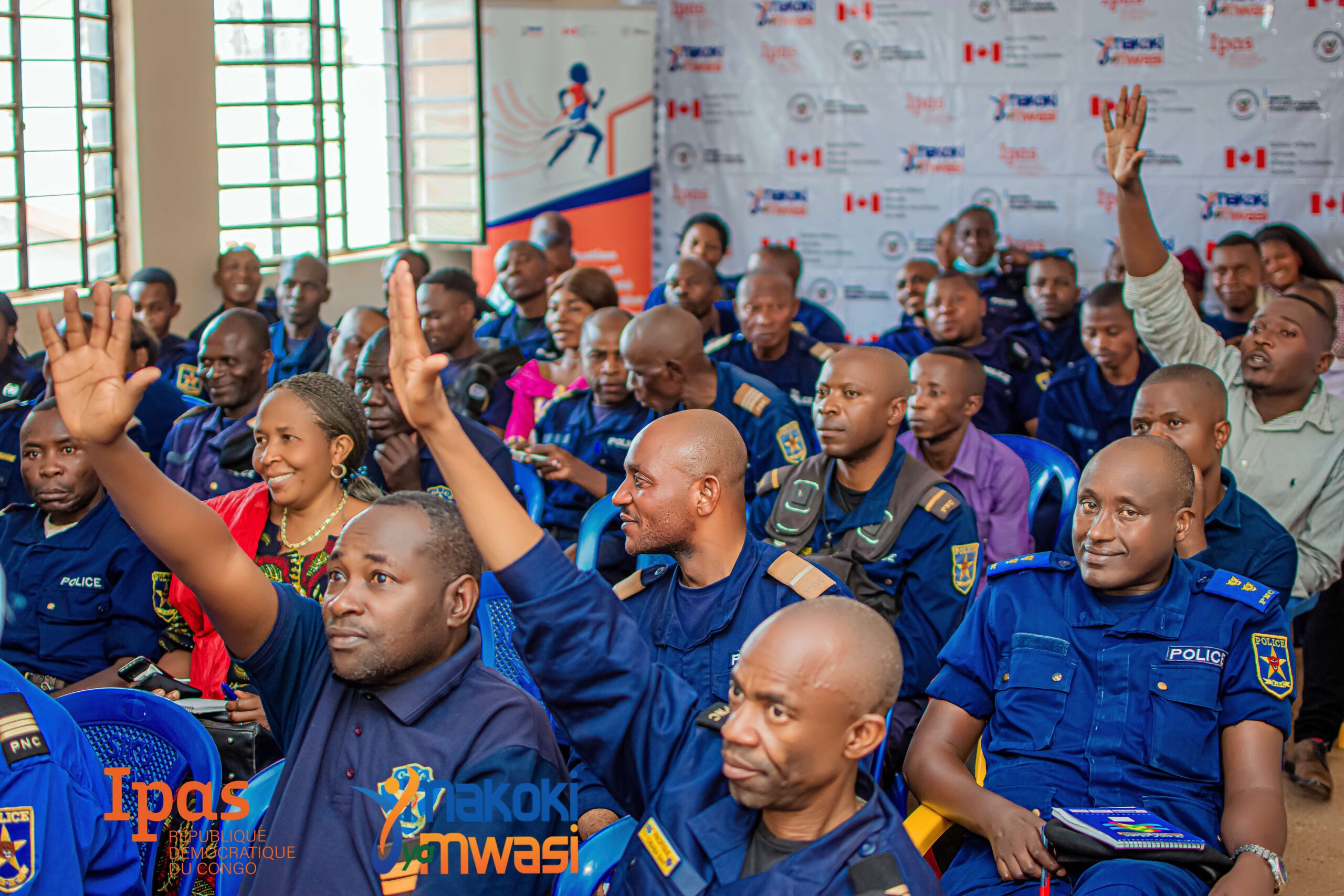 A group of police officers and civilians sit in a classroom, some raising their hands to participate. Logos for Ipas and Makoki Mwasi are visible in the foreground, with banners and logos displayed on the back wall. A group of police officers and civilians sit in a classroom, some raising their hands to participate. Logos for Ipas and Makoki Mwasi are visible in the foreground, with banners and logos displayed on the back wall.