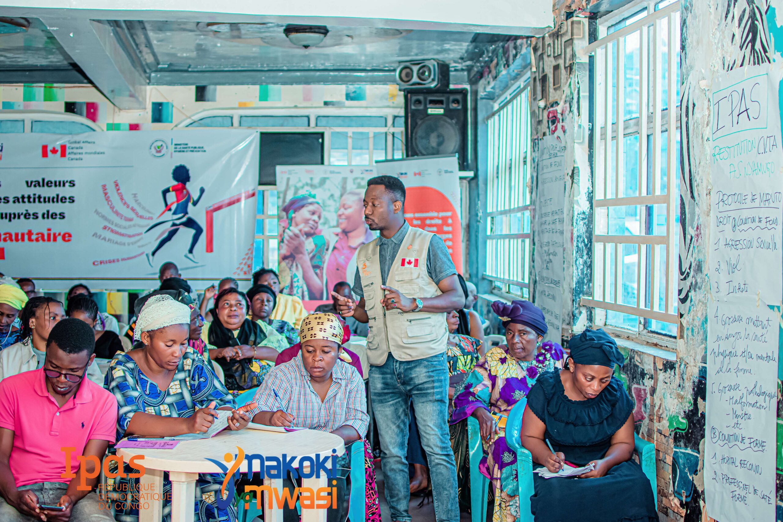 A man wearing a Canada-branded vest speaks to a diverse group of people seated and taking notes in a colorful, poster-filled classroom during a workshop or training session. A man wearing a Canada-branded vest speaks to a diverse group of people seated and taking notes in a colorful, poster-filled classroom during a workshop or training session.
