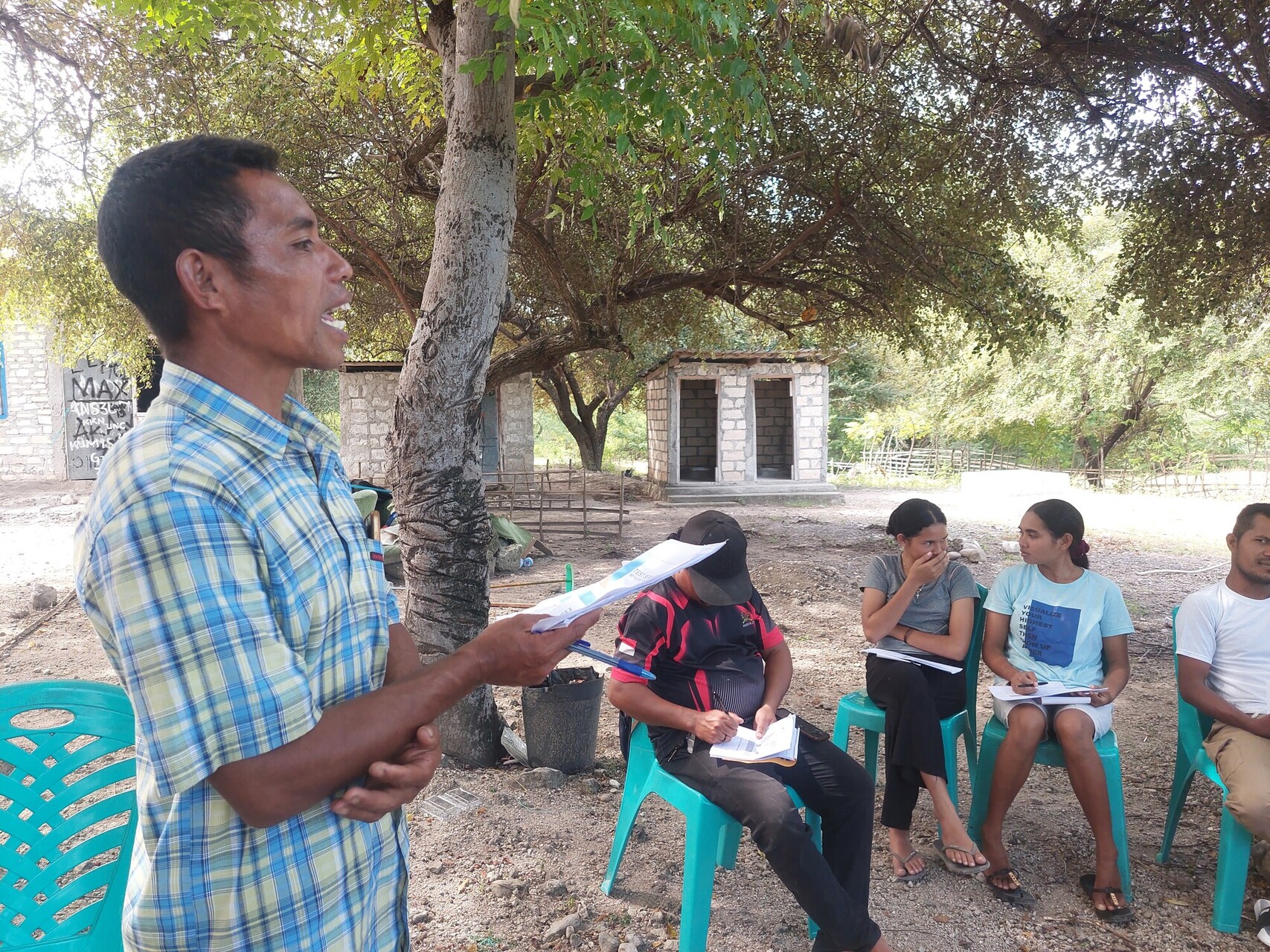 A man stands and speaks outdoors, holding papers, while four people sit and listen on blue chairs under trees, taking notes. There is a building in the background.