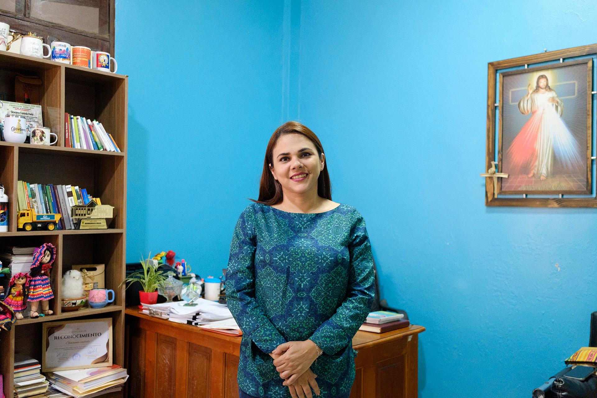 A woman in a blue patterned dress stands smiling in an office with blue walls, a bookshelf with mugs and books, a wooden desk with papers, and a framed religious image hanging on the wall. A woman in a blue patterned dress stands smiling in an office with blue walls, a bookshelf with mugs and books, a wooden desk with papers, and a framed religious image hanging on the wall.