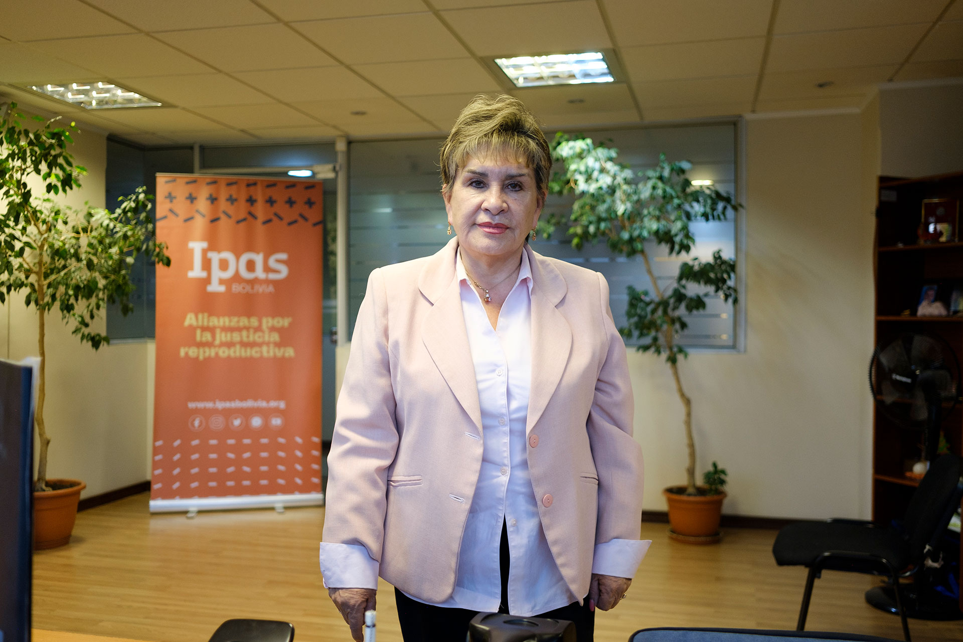 A person with short hair, wearing a light pink blazer and white shirt, stands in an office. Behind them is an orange banner with the words "Ipas Bolivia, Alianzas por la justicia reproductiva.