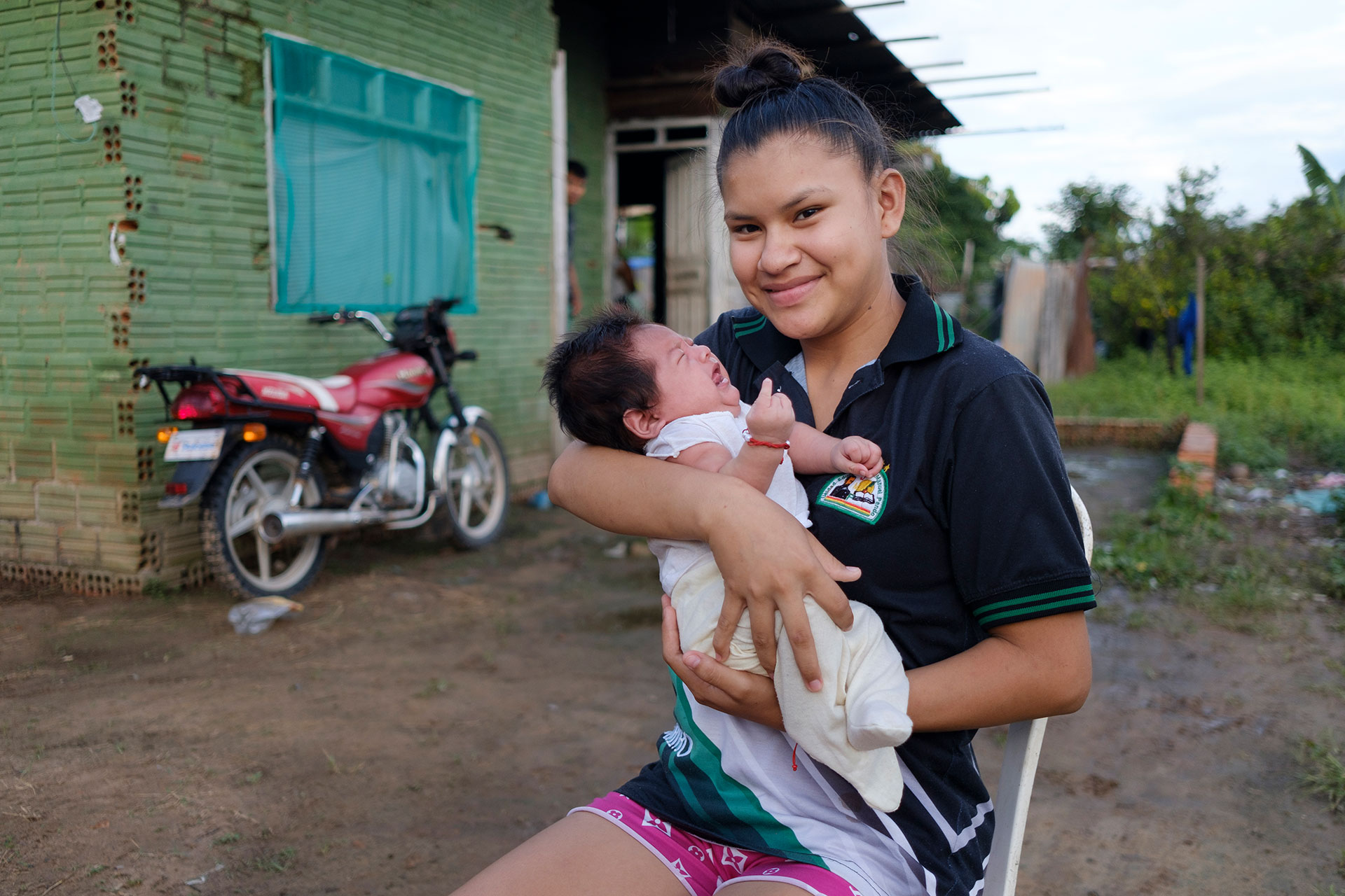 A young woman sits outside on a chair, smiling at the camera while holding a baby in her arms. A red motorcycle is parked nearby, and a green brick building is in the background. The scene appears calm and relaxed.
