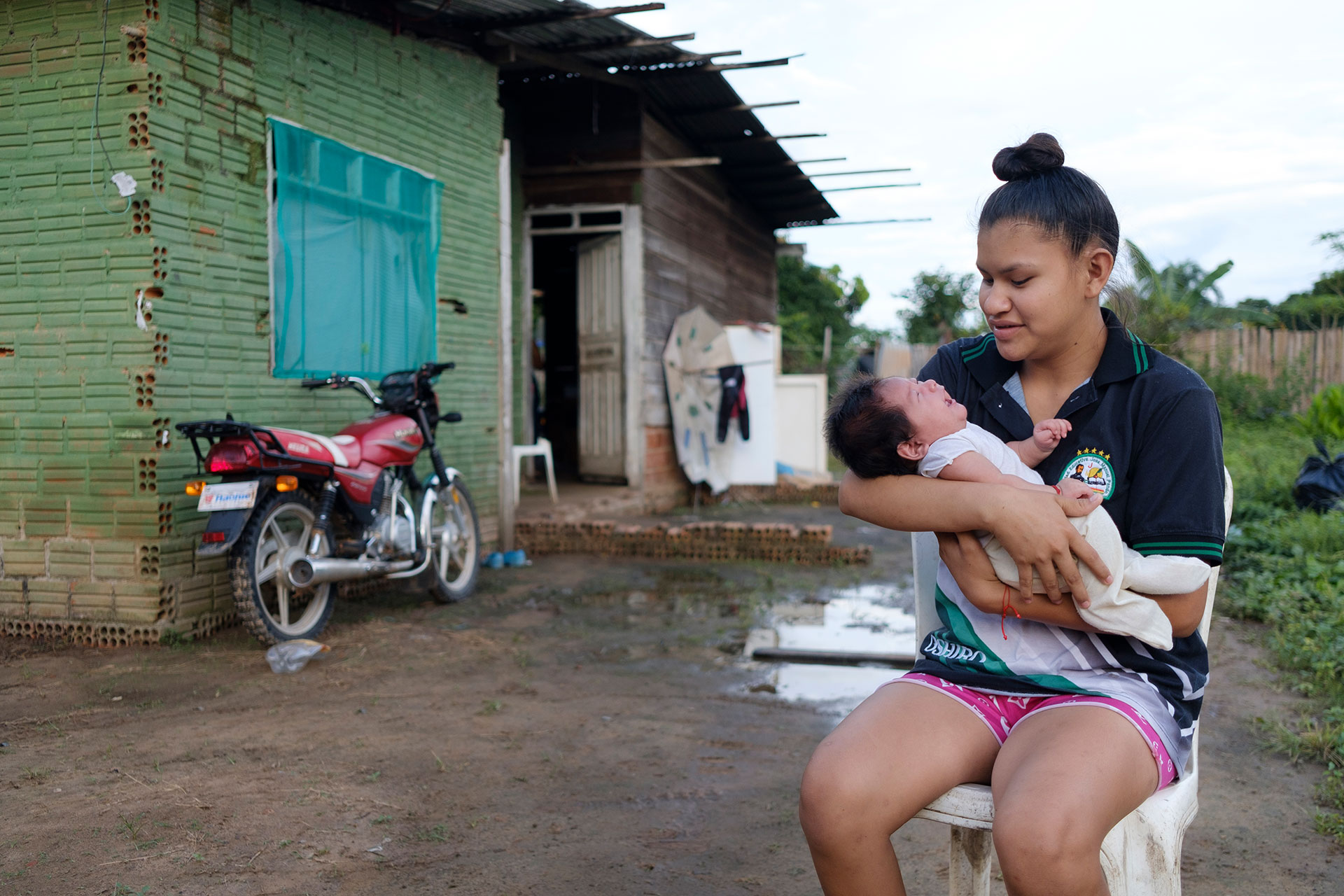 A young woman sits on a stool outside a rustic house, cradling a newborn baby. A red motorcycle is parked nearby, and laundry hangs by the doorway. The ground is muddy and the area is surrounded by greenery.