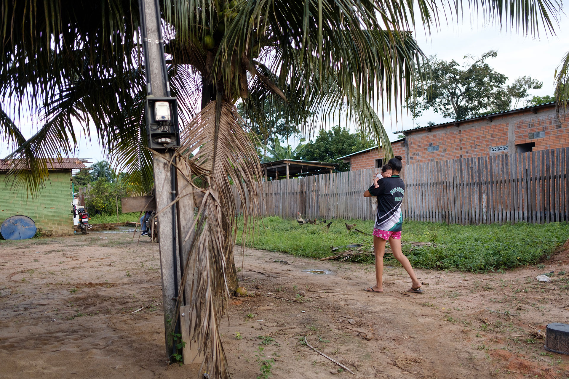 A person wearing shorts and a dark shirt carries a small child while walking barefoot on sandy ground near a palm tree, with wooden fences, houses, and greenery in the background. A person wearing shorts and a dark shirt carries a small child while walking barefoot on sandy ground near a palm tree, with wooden fences, houses, and greenery in the background.