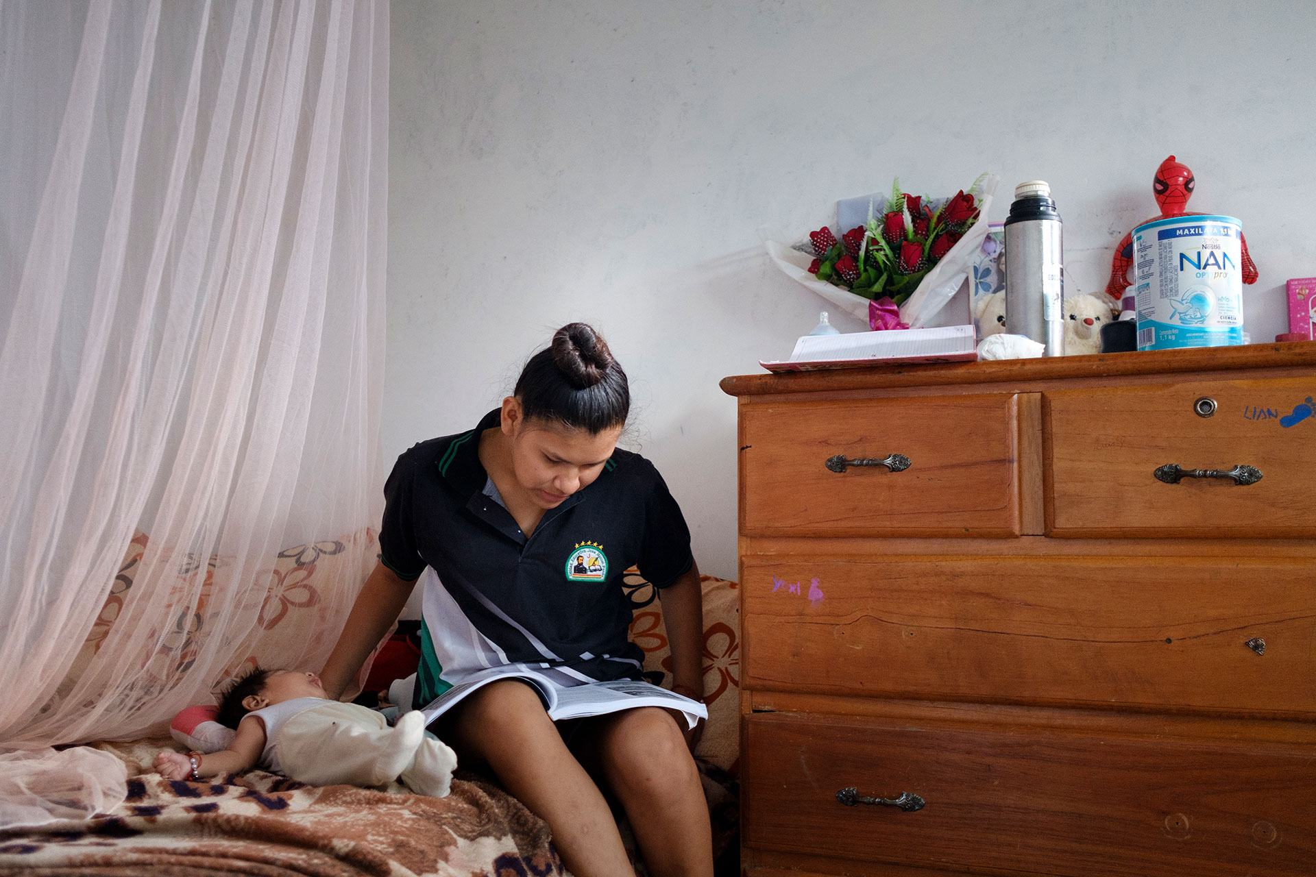 A woman sits on a bed reading a book, with a baby lying beside her. Next to her, a wooden dresser holds a bouquet, a thermos, a can of infant formula, and a few small items against a white wall. A woman sits on a bed reading a book, with a baby lying beside her. Next to her, a wooden dresser holds a bouquet, a thermos, a can of infant formula, and a few small items against a white wall.