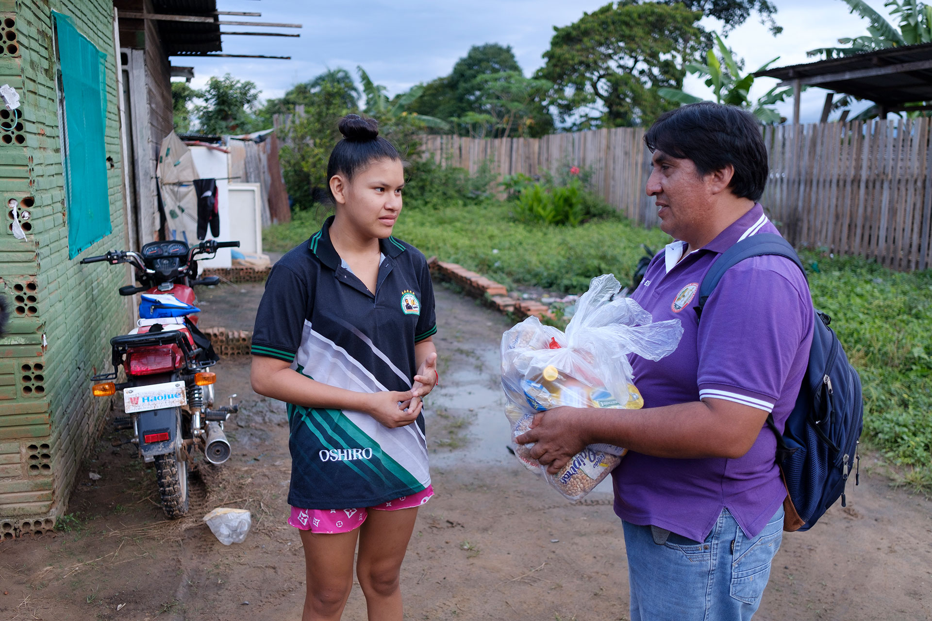 A man in a purple shirt hands a clear bag of groceries to a young woman outside a house, with a motorcycle and greenery visible in the background. A man in a purple shirt hands a clear bag of groceries to a young woman outside a house, with a motorcycle and greenery visible in the background.