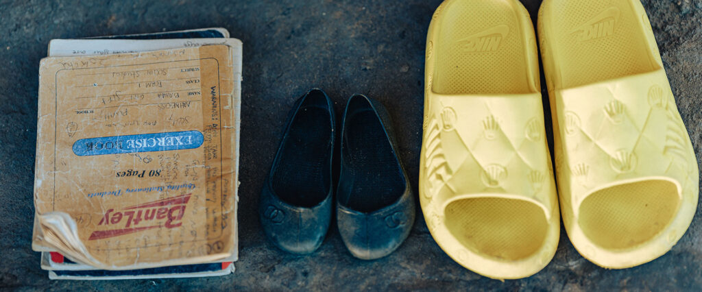 A pair of worn black shoes, yellow Nike sandals, and a stack of exercise books with handwritten notes are neatly placed on a rough, dark surface.