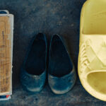A pair of worn black shoes, yellow Nike sandals, and a stack of exercise books with handwritten notes are neatly placed on a rough, dark surface.