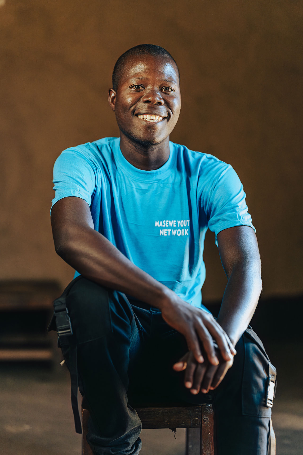 A smiling young man sits on a wooden stool, wearing a bright blue t-shirt with the words "MASEVE YOUTH NETWORK" printed on it, in a warmly lit indoor setting with a plain brown background. A smiling young man sits on a wooden stool, wearing a bright blue t-shirt with the words "MASEVE YOUTH NETWORK" printed on it, in a warmly lit indoor setting with a plain brown background.