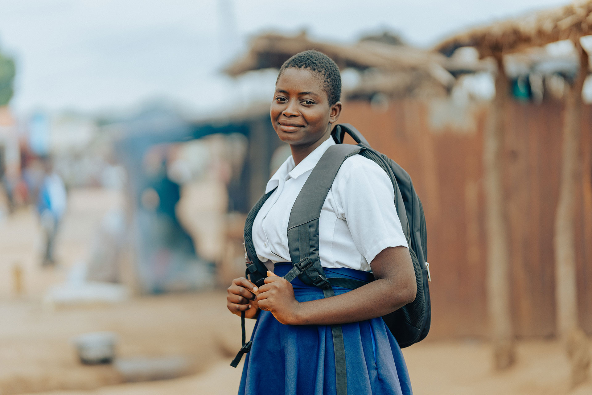 A young girl in a school uniform and backpack smiles while standing outdoors in a rural area, with wooden structures and a blurred background behind her.