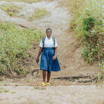 A woman wearing a blue skirt, white blouse, and yellow sandals stands on a dirt path surrounded by tall green grass, with a backpack on her shoulders.