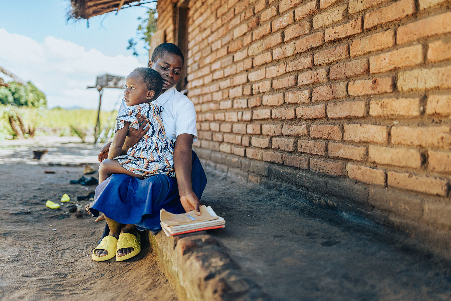 A young girl in a school uniform sits on a low brick wall, holding a small child on her lap and reading from a book, outside a rustic building on a sunny day. A young girl in a school uniform sits on a low brick wall, holding a small child on her lap and reading from a book, outside a rustic building on a sunny day.