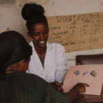 A woman in a white coat shows educational materials with diagrams to another woman across a table in a modest, worn classroom with charts and notes on the wall.