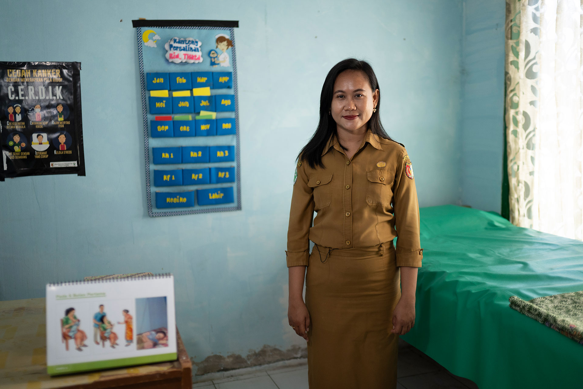 A woman in a tan uniform stands in a classroom with light blue walls, educational posters, a table with a calendar, and a green-covered bed near a window with sheer curtains.