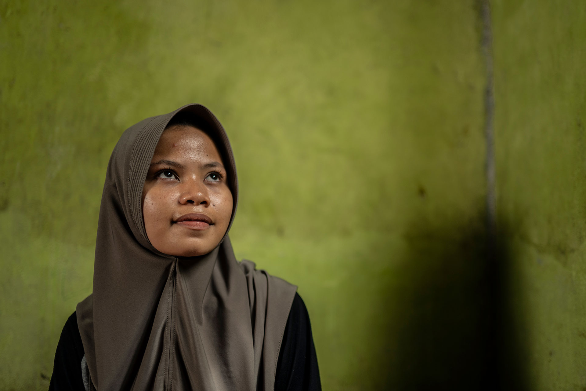 A young woman wearing a taupe hijab looks upward with a thoughtful expression, standing against a textured green wall with a shadow cast to the right.