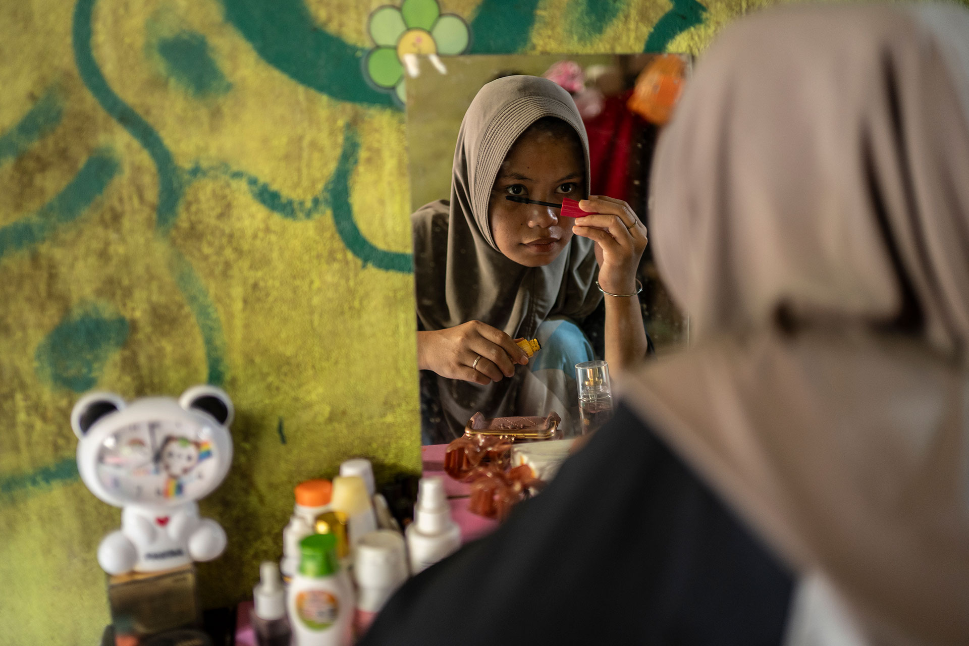 A girl in a headscarf applies makeup while looking in a mirror on a cluttered vanity with bottles, a clock, and a toy bear, against a yellow-green wall with painted designs.