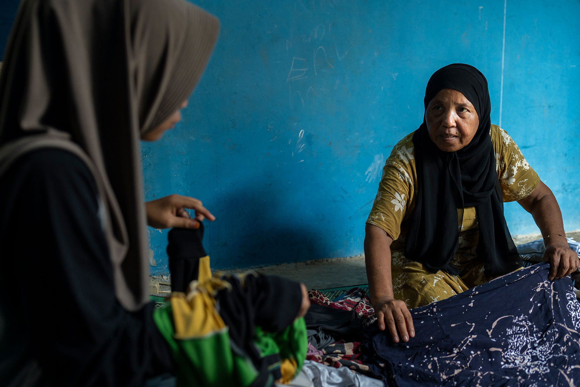 Two women wearing headscarves sit indoors against a blue wall, sorting and folding clothes laid out on the floor. One woman wears a yellow patterned dress and looks toward the other, who holds a stack of clothes.