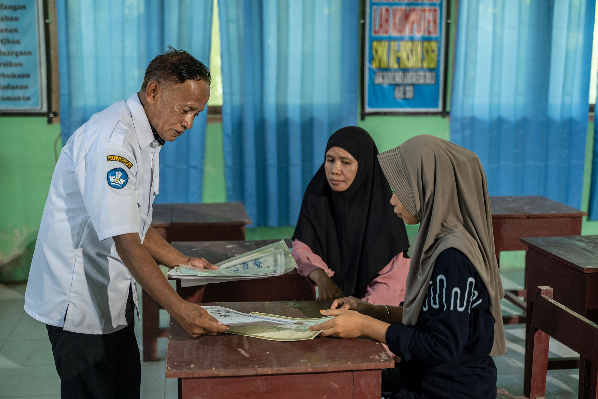 A man in a white shirt hands documents to a seated young woman in a hijab, while another woman in a hijab sits beside her in a classroom with blue curtains and educational posters on the walls.