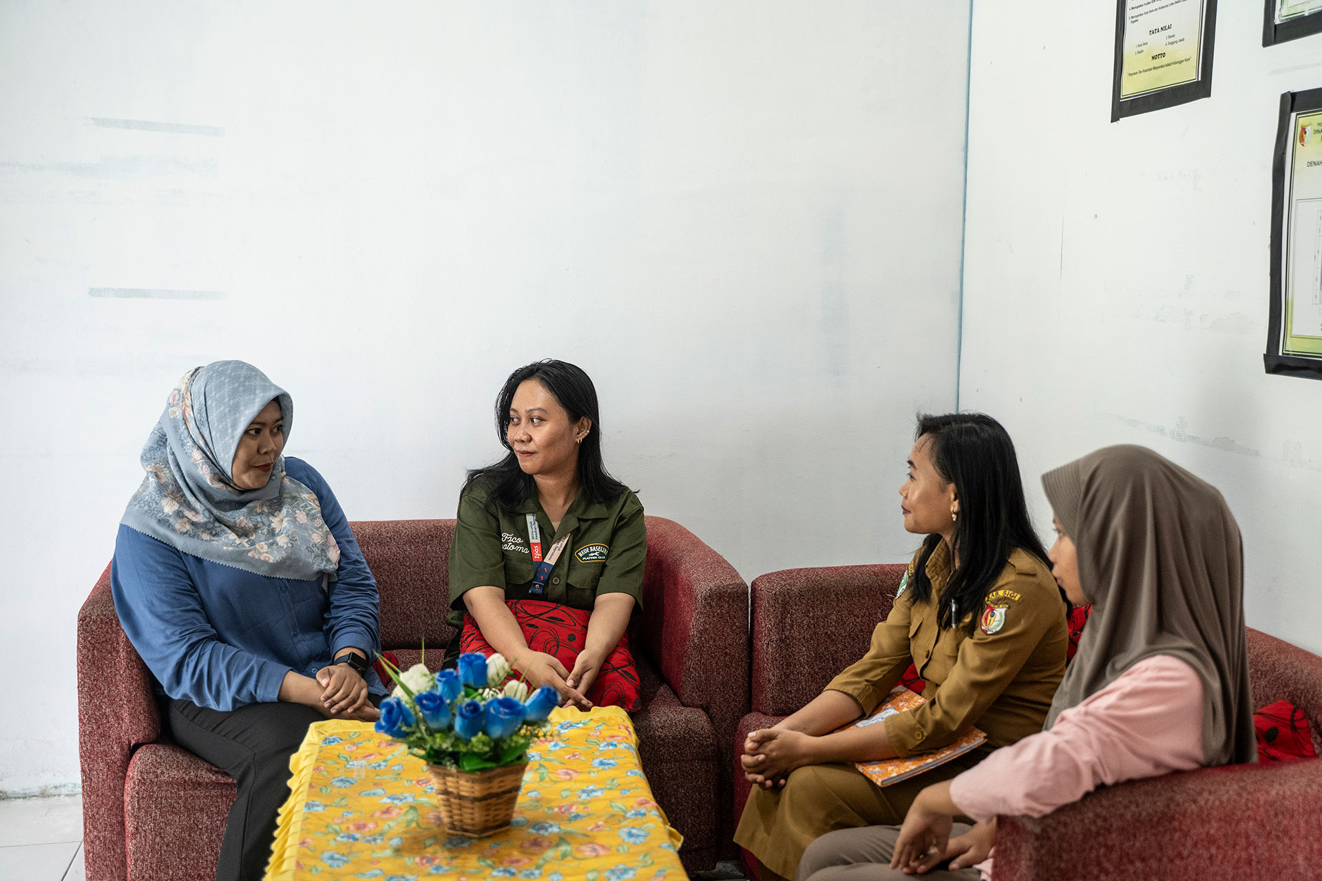 Four women are sitting on red sofas around a table with a yellow cloth and a basket of blue flowers, having a conversation in a brightly lit room with framed certificates on the wall. Four women are sitting on red sofas around a table with a yellow cloth and a basket of blue flowers, having a conversation in a brightly lit room with framed certificates on the wall.