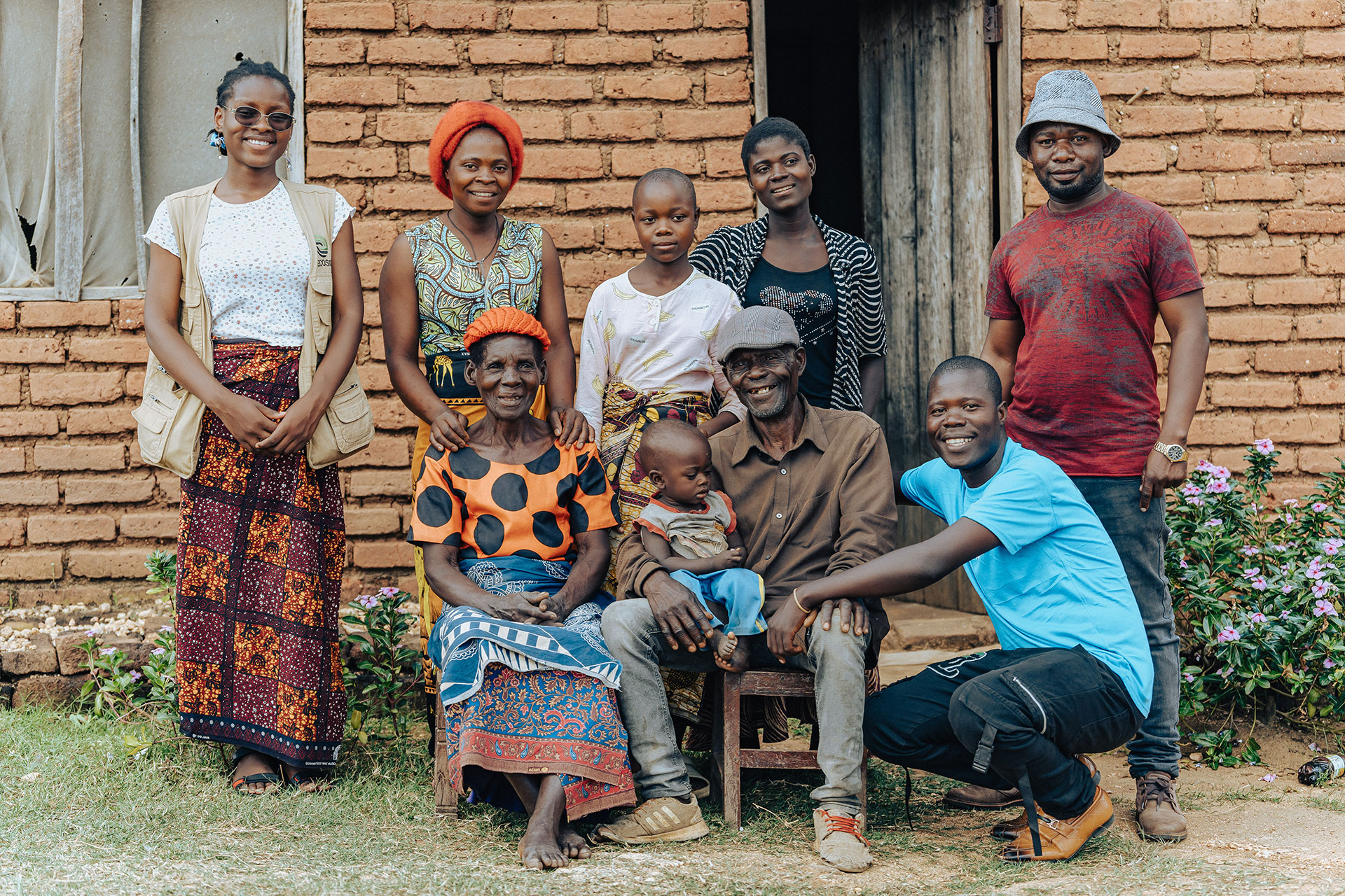 A group of nine people, including adults and children, pose and smile outside a brick house. Two people are seated, one holding a toddler, while the others stand or crouch around them, all dressed in colorful clothing.