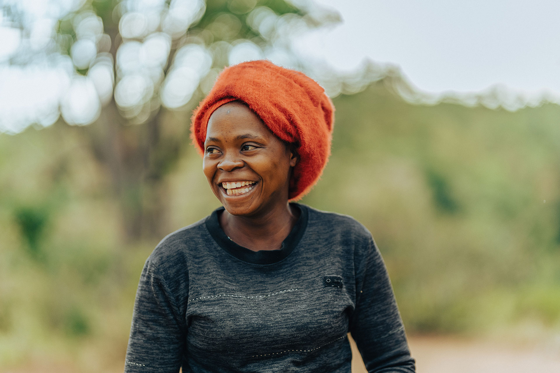 A woman wearing a bright red headwrap and a dark sweater smiles warmly outdoors, with green trees and a soft, blurred background behind her.