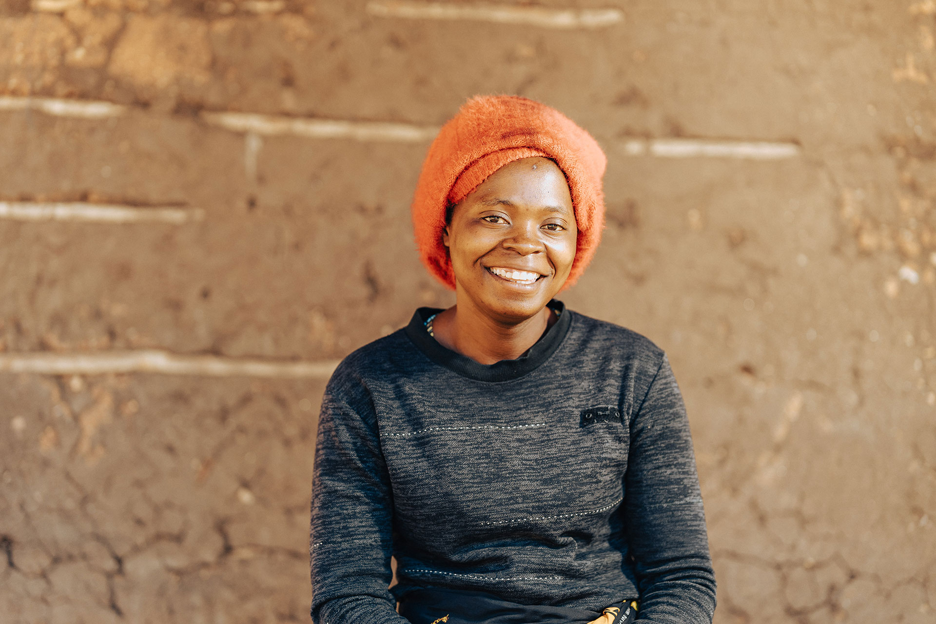 A woman wearing a bright orange headscarf and a dark sweater smiles while sitting in front of a textured, brown mud wall.