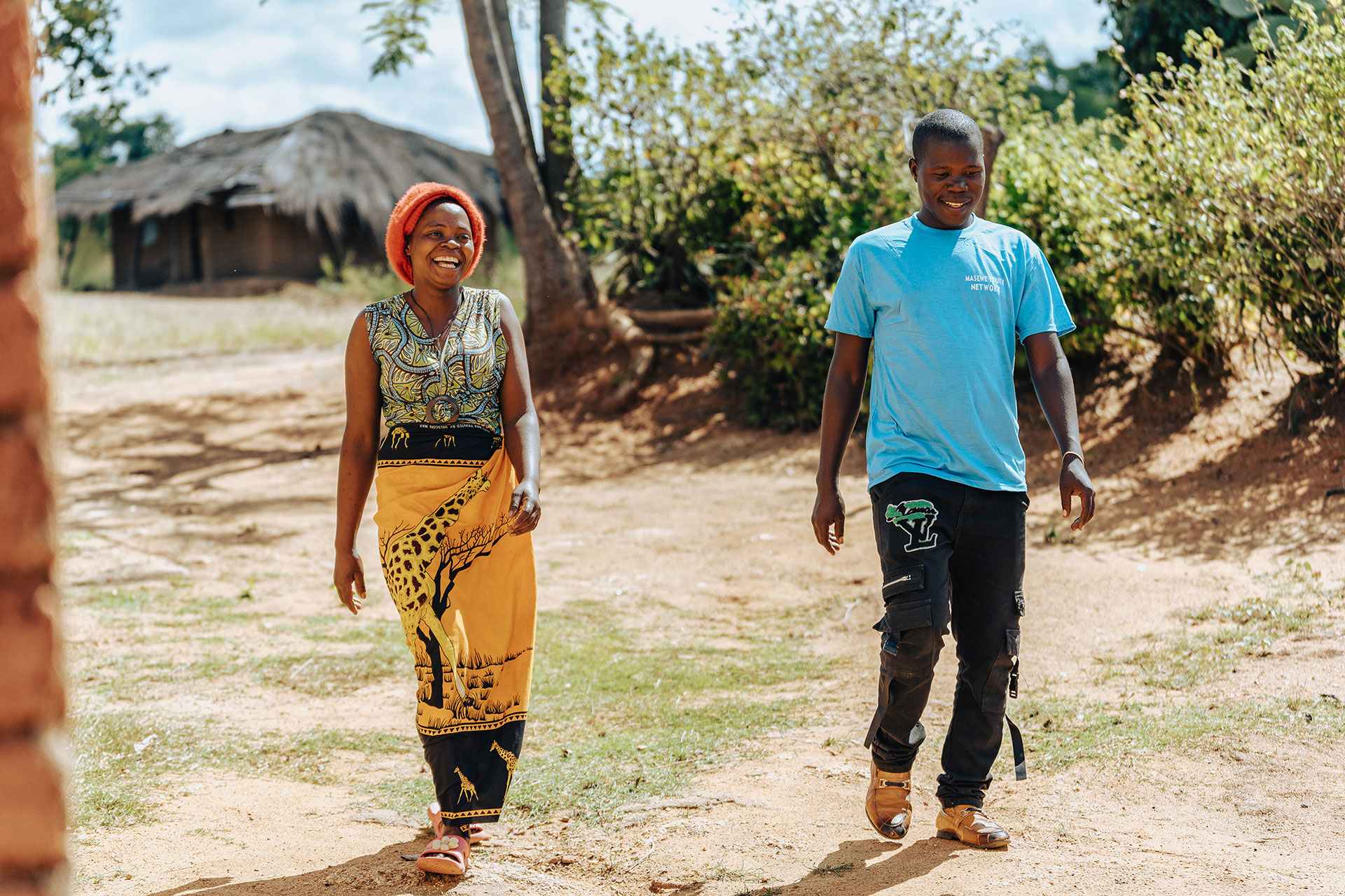 A smiling woman in a colorful dress and red headscarf walks beside a man in a blue shirt and dark pants on a dirt path, with trees and a thatched-roof hut in the background. A smiling woman in a colorful dress and red headscarf walks beside a man in a blue shirt and dark pants on a dirt path, with trees and a thatched-roof hut in the background.