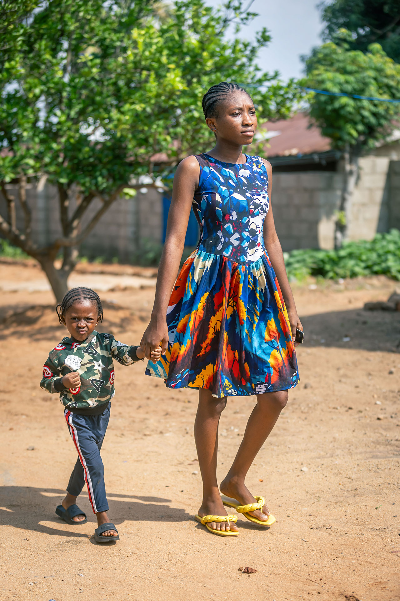 A woman in a colorful dress and yellow sandals holds hands with a young child in a black and green outfit as they walk outdoors on a sandy path, with green trees and a wall in the background. A woman in a colorful dress and yellow sandals holds hands with a young child in a black and green outfit as they walk outdoors on a sandy path, with green trees and a wall in the background.