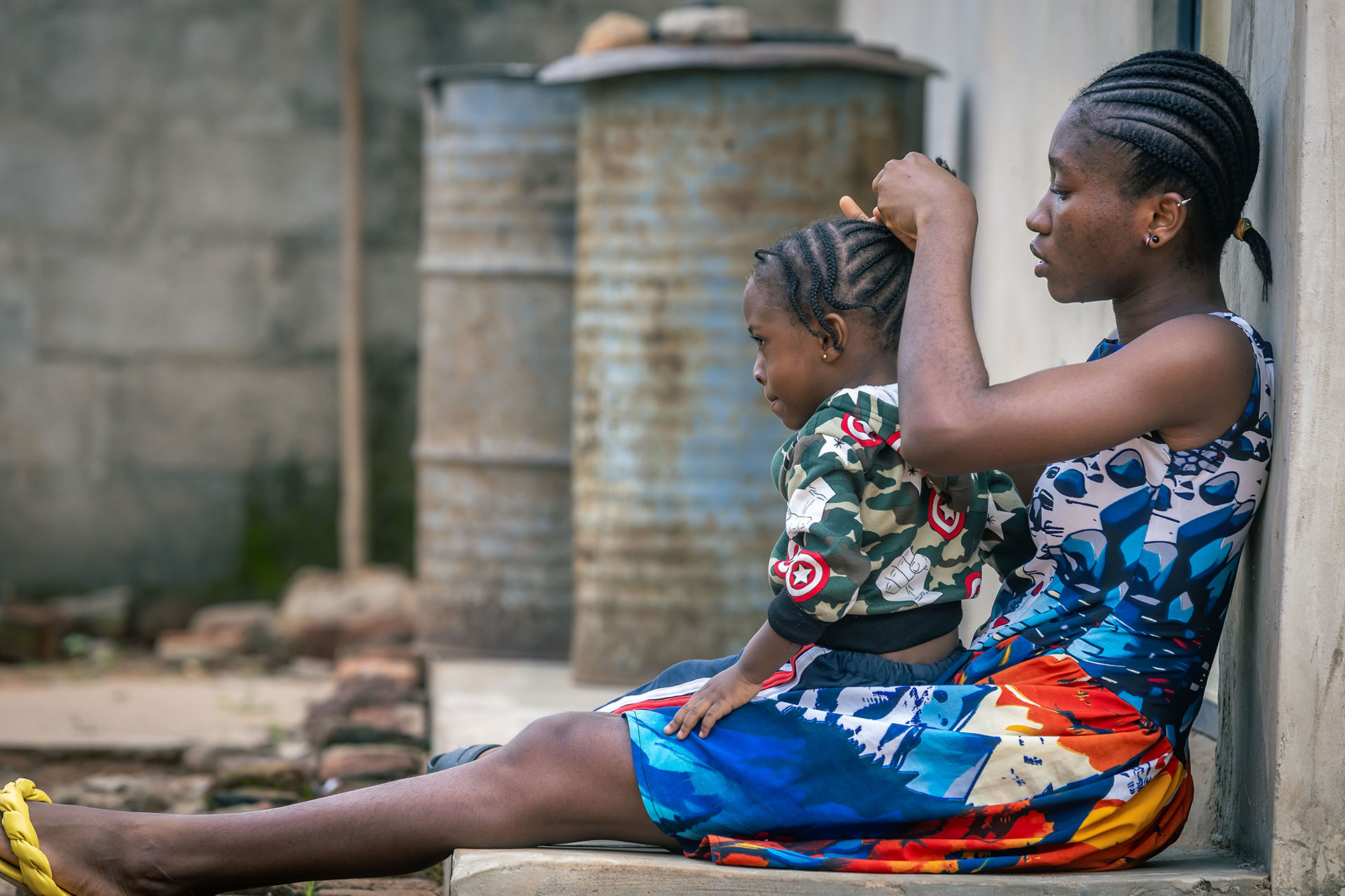 A woman sits on a concrete step outside, braiding a young girl’s hair. Both have braided styles, and the child wears a patterned jacket. Two metal barrels and a rough wall are visible in the background. A woman sits on a concrete step outside, braiding a young girl’s hair. Both have braided styles, and the child wears a patterned jacket. Two metal barrels and a rough wall are visible in the background.