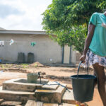 A young girl carries a black bucket of water from a well in a sunlit, dusty yard with trees, a rope with clothes, and a house in the background.