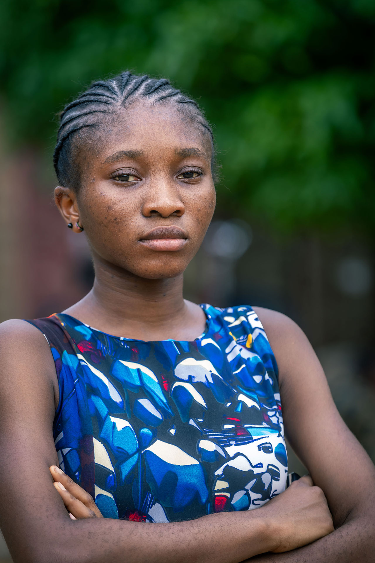 A young woman with braided hair and a serious expression stands outdoors with her arms crossed, wearing a colorful, blue-patterned sleeveless top. Green foliage is blurred in the background. A young woman with braided hair and a serious expression stands outdoors with her arms crossed, wearing a colorful, blue-patterned sleeveless top. Green foliage is blurred in the background.
