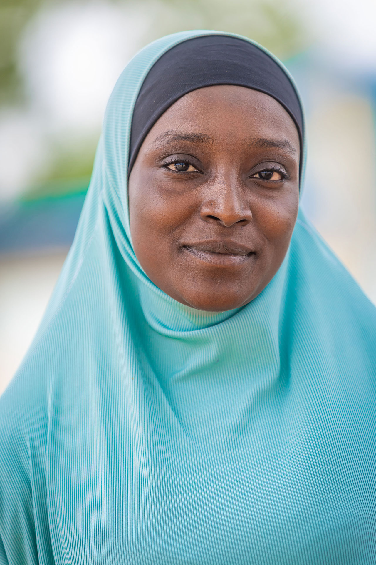 A woman wearing a light blue hijab and a black undercap looks at the camera with a gentle expression. The background is softly blurred with hints of greenery and sky. A woman wearing a light blue hijab and a black undercap looks at the camera with a gentle expression. The background is softly blurred with hints of greenery and sky.